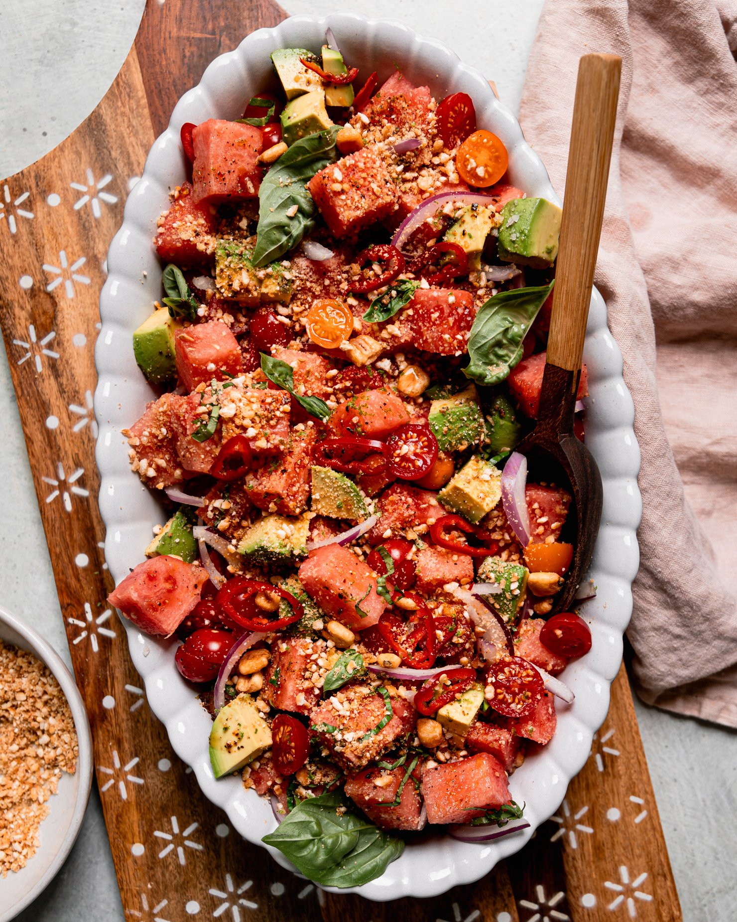 An overhead shot shows an oval platter filled with watermelon salad that also features avocado, sliced red onion, halved cherry tomatoes, and finely crushed salted corn nuts. Fresh basil leaves garnish the top and a wooden serving spoon is sticking out of the dish.