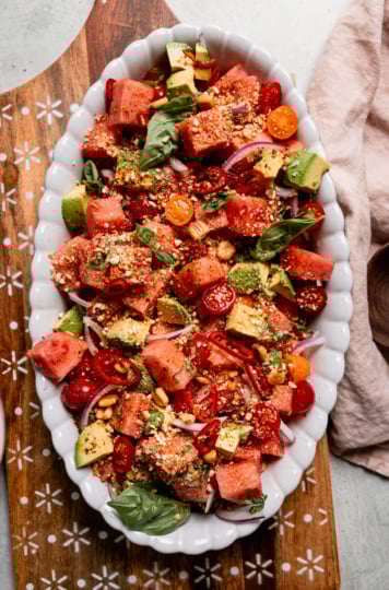 An overhead shot shows an oval platter filled with watermelon salad that also features avocado, sliced red onion, halved cherry tomatoes, and finely crushed salted corn nuts. Fresh basil leaves garnish the top and a napkin plus wooden serving spoon are seen to the side.