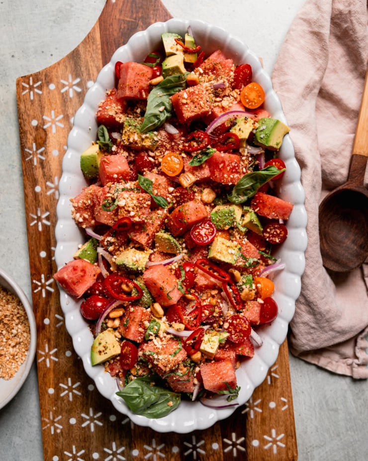 An overhead shot shows an oval platter filled with watermelon salad that also features avocado, sliced red onion, halved cherry tomatoes, and finely crushed salted corn nuts. Fresh basil leaves garnish the top and a napkin plus wooden serving spoon are seen to the side.