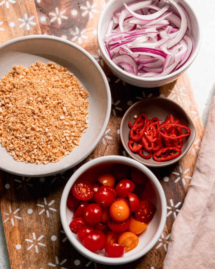 An overhead shot shows prepped ingredients in bowls: sliced red onions, sliced fresno chilies, halved cherry tomatoes, and ground up salted corn nuts.