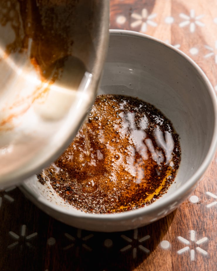 An overhead shot shows sizzling hot oil being poured into a bowl with ground chilies and ground black pepper.