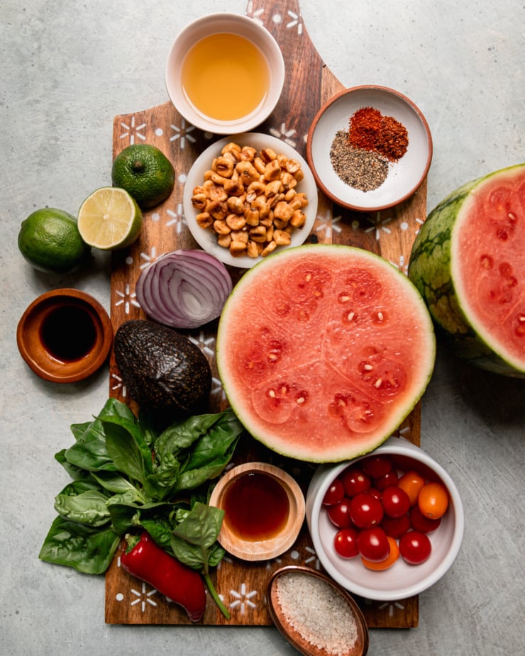 An overhead shot shows ingredients for a salad: ground chilies, corn nuts, watermelon, cherry tomatoes, salt, maple syrup, Fresno chili, basil, avocado, red onion, Tamari, limes, and avocado oil.
