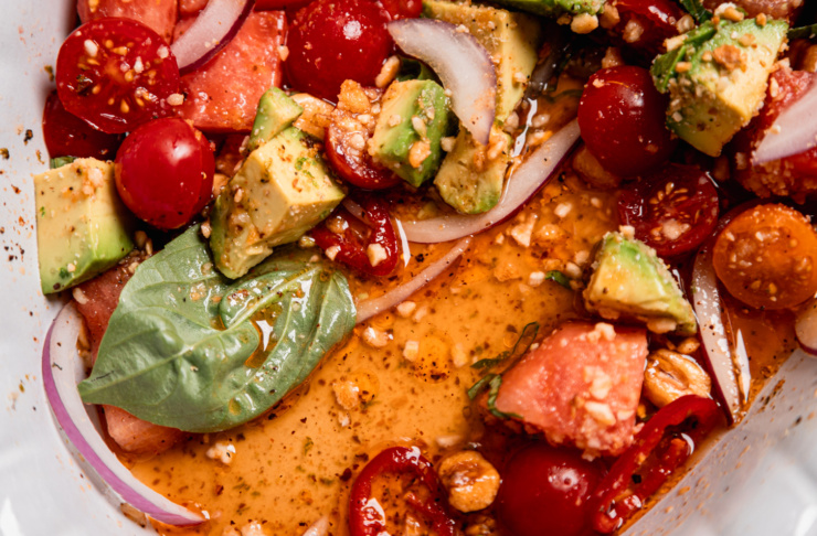An up close overhead shot shows the remains of a watermelon salad with basil, Fresno chili, avocado, basil, red onion, cherry tomatoes, and finely ground corn nuts. There is a pool of dressing in the bottom of the dish.