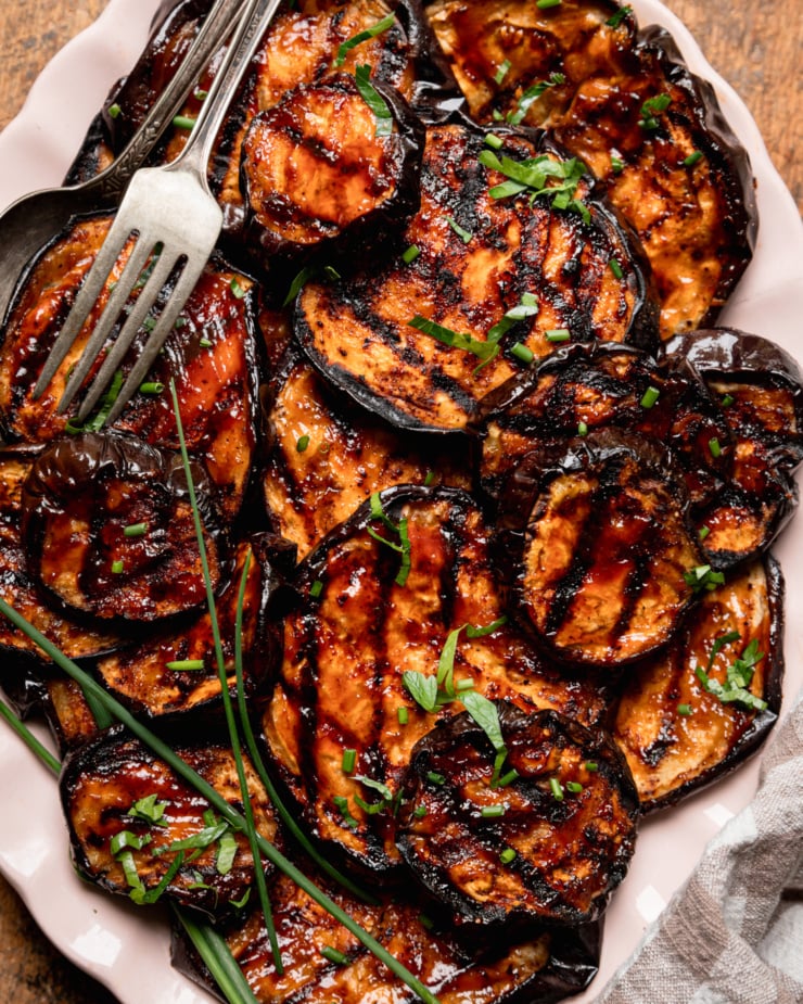An up close, overhead shot shows grilled eggplant slices brushed with barbecue sauce on an oval platter. The slices are garnished with chopped herbs.