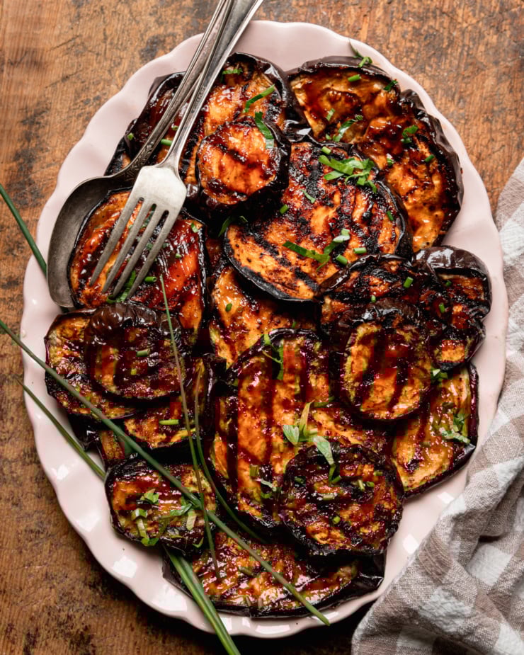 An overhead shot shows grilled eggplant slices brushed with barbecue sauce on an oval platter. The slices are garnished with chopped herbs.