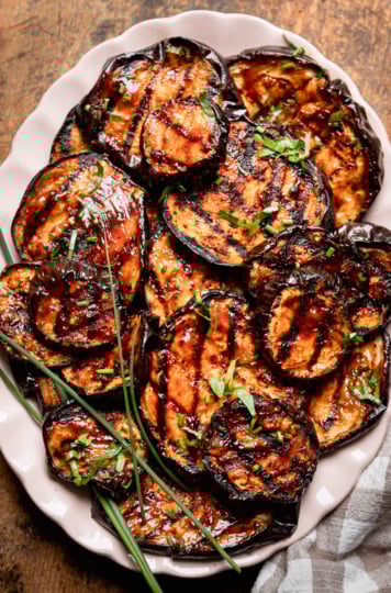 An overhead shot shows grilled eggplant slices brushed with barbecue sauce on an oval platter. The slices are garnished with chopped herbs.