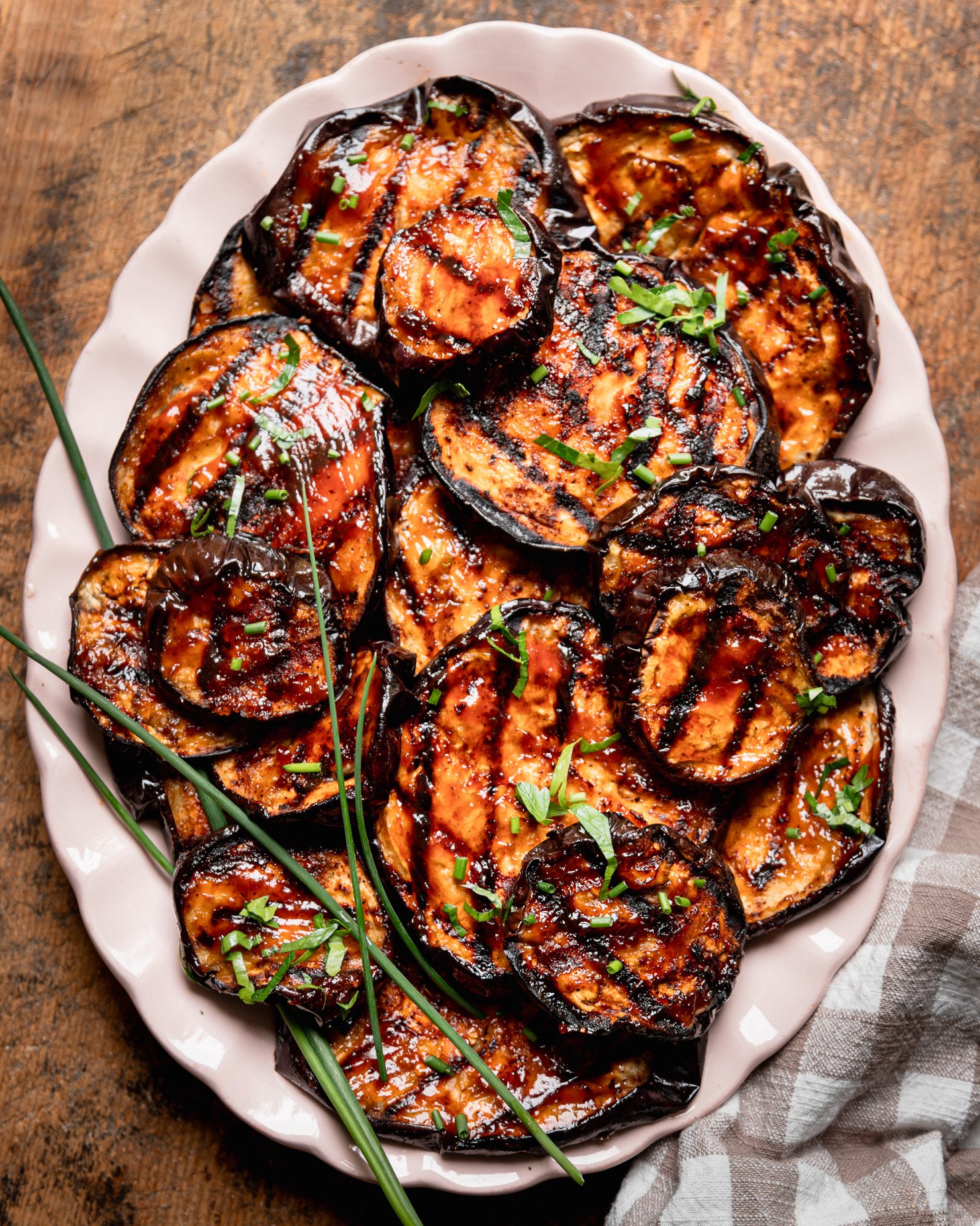 An overhead shot shows grilled eggplant slices brushed with barbecue sauce on an oval platter. The slices are garnished with chopped herbs.