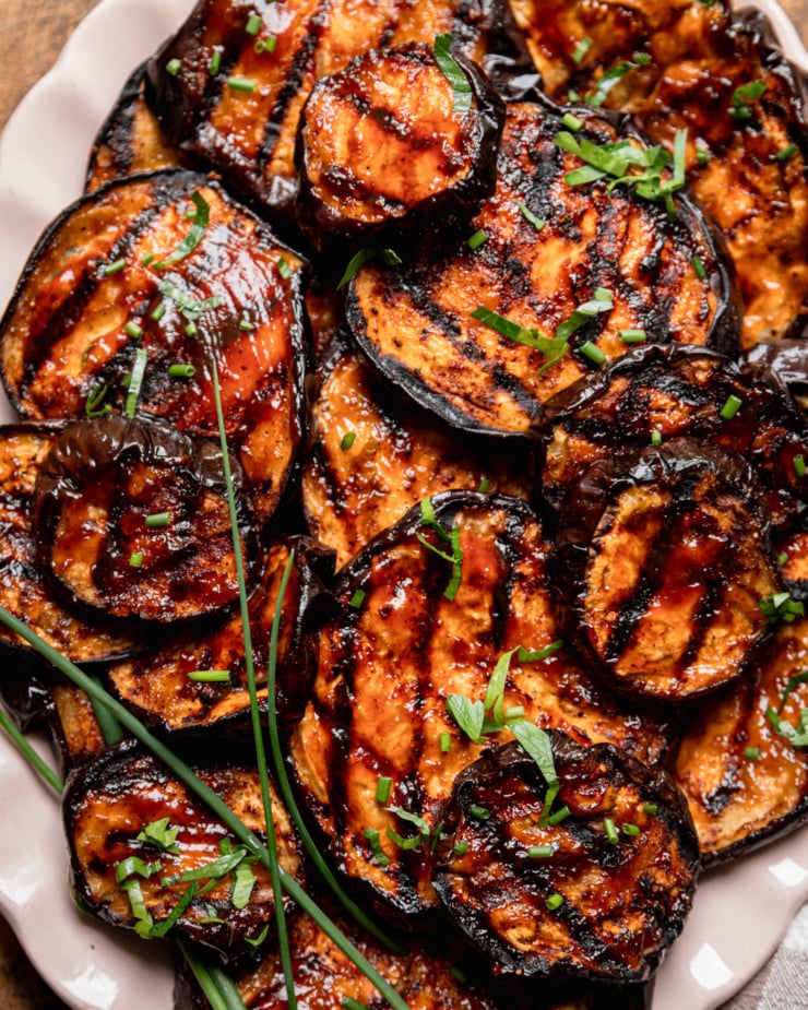 An up close, overhead shot shows grilled eggplant slices brushed with barbecue sauce on an oval platter. The slices are garnished with chopped herbs.