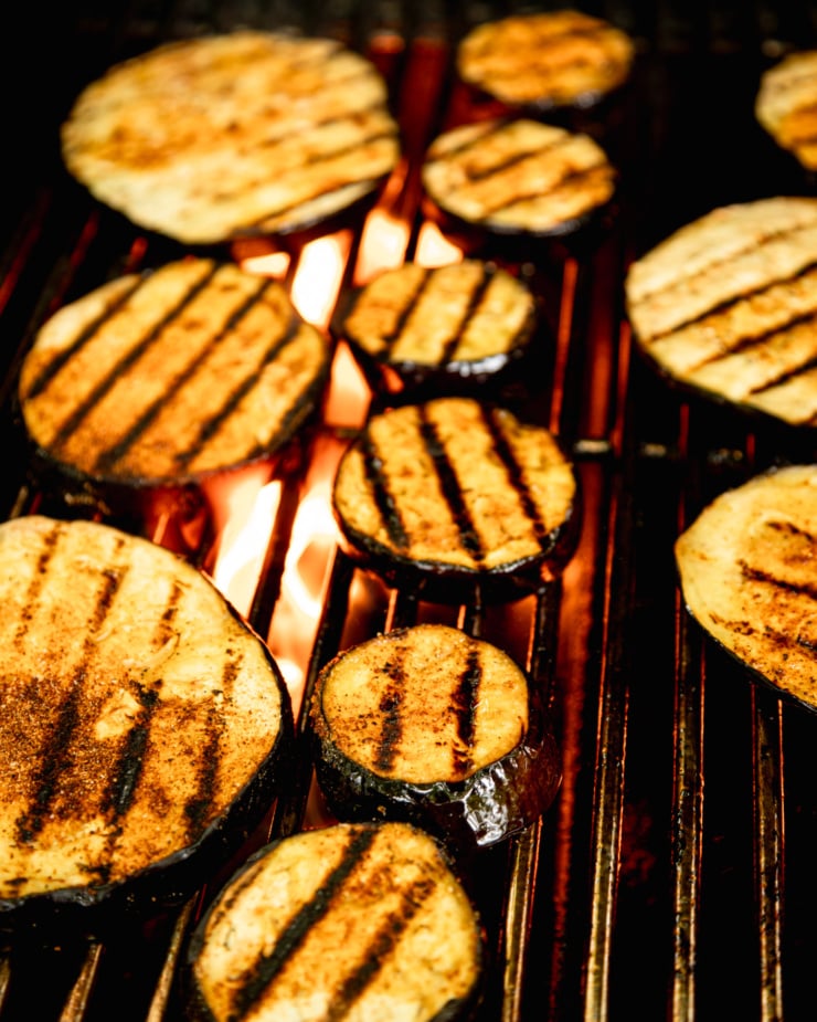 An overhead shot shows slices of eggplant with char marks on a BBQ. There are flames kicking up under the grates.