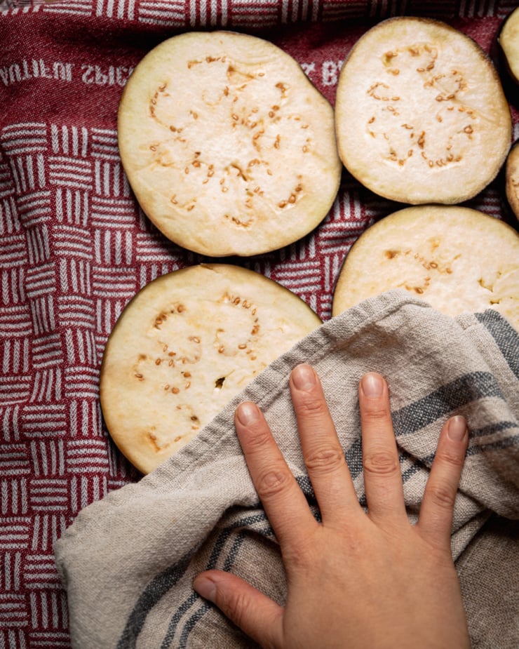 An overhead shot shows a hand using a towel to dry off some slices of eggplant.