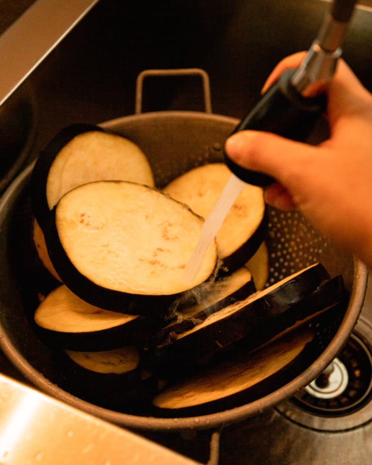 An overhead shot shows slices of eggplant in a colander int he sink. A hand is using the hose of the faucet to rinse them off.
