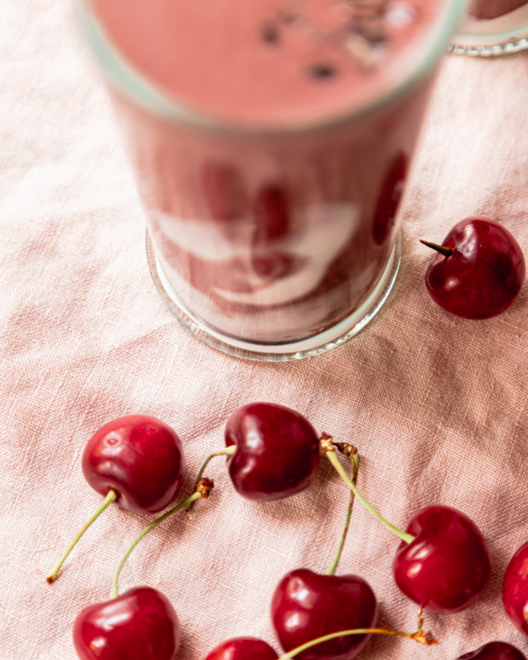 An overhead shot shows a blurry glass filled with a smoothie and some fresh cherries on a linen napkin.