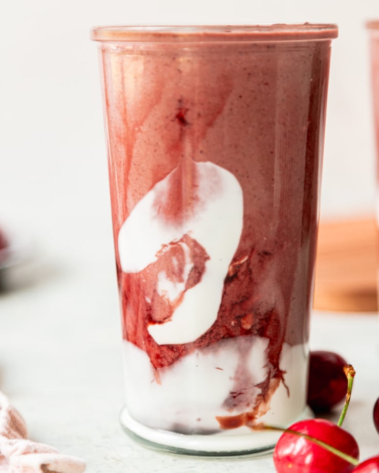 An up close, head-on shot shows a glass of chocolate cherry smoothie. The glass is swirled with coconut milk yogurt and tart cherry juice concentrate. Fresh cherries are seen in the background.