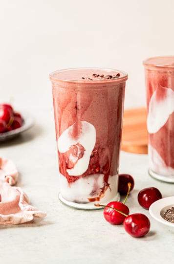 A slight 3/4 angle shot shows a glass of chocolate cherry smoothie. The glass is swirled with coconut milk yogurt and tart cherry juice concentrate. Fresh cherries are seen in the background.