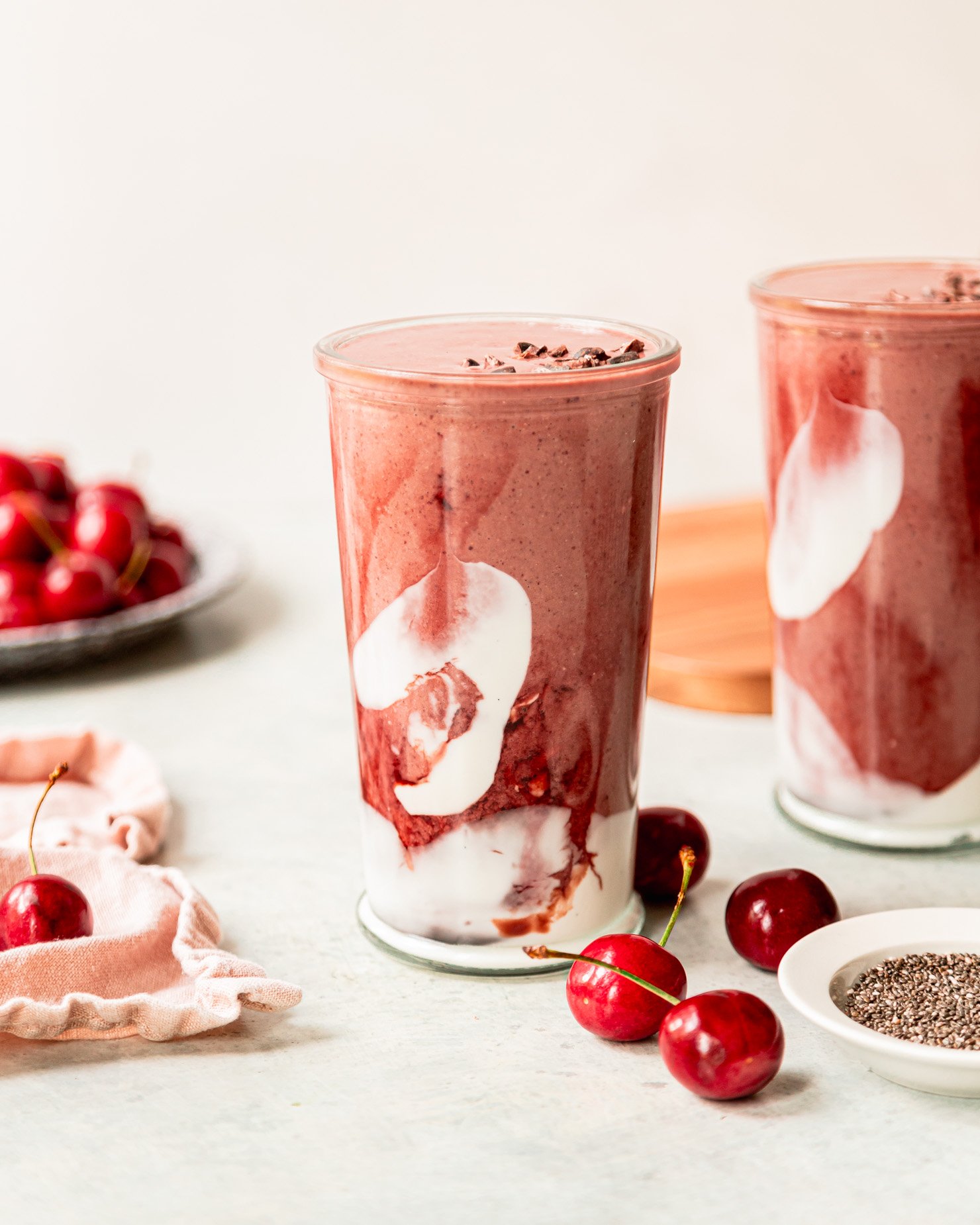 A slight 3/4 angle shot shows a glass of chocolate cherry smoothie. The glass is swirled with coconut milk yogurt and tart cherry juice concentrate. Fresh cherries are seen in the background.