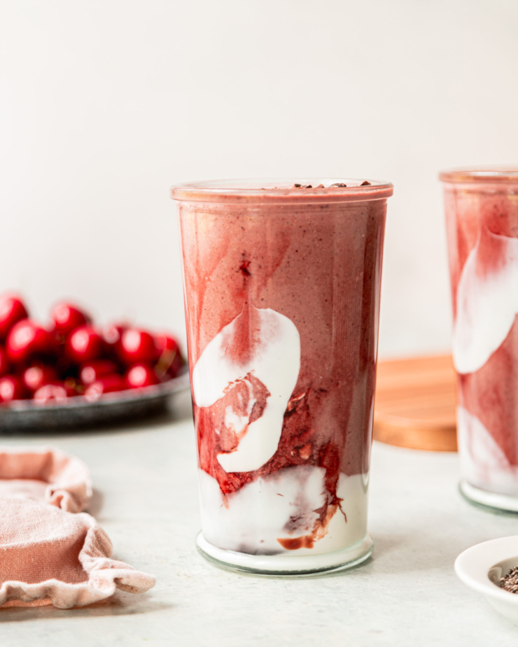 A head-on shot shows a glass of chocolate cherry smoothie. The glass is swirled with coconut milk yogurt and tart cherry juice concentrate. Fresh cherries are seen in the background.