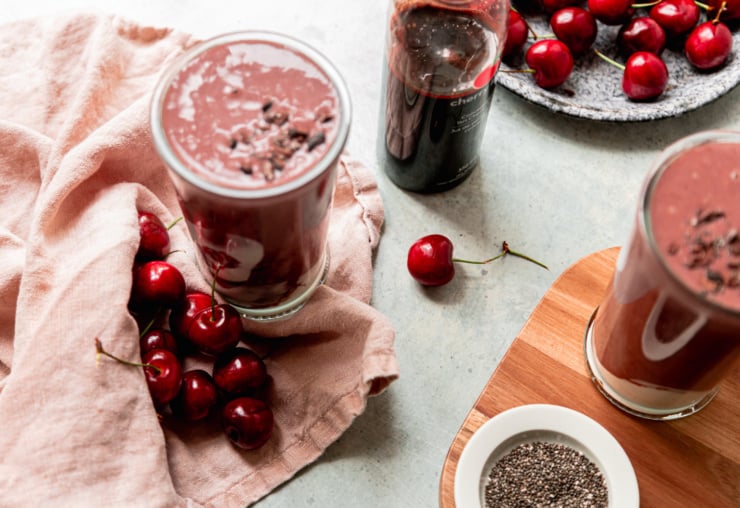 An overhead shot shows two glasses of chocolate cherry smoothie with fresh cherries, tart cherry concentrate juice, and a pink napkin all nearby.