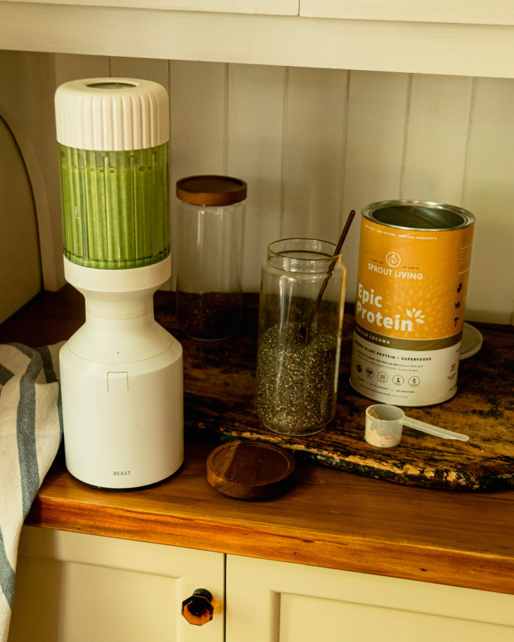 A slight 3/4 angle shot shows a blender on the kitchen counter with a green smoothie blended up inside. On the counter are jars of chia seeds, flax seeds, and vegan protein powder.