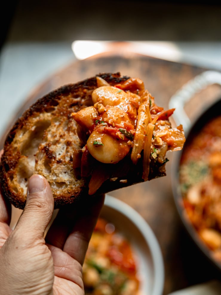 An overhead shot shows a hand holding a crusty piece of bread with a saucy butter bean mixture piled on top.