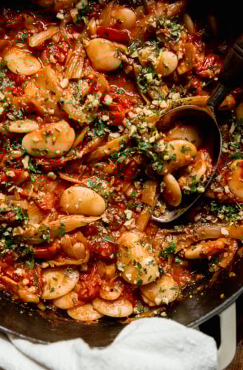 An up close, overhead shot shows a caramelized fennel, tomato and butter bean skillet garnished with chopped parsley and walnuts. A serving spoon is sticking out of the pan.