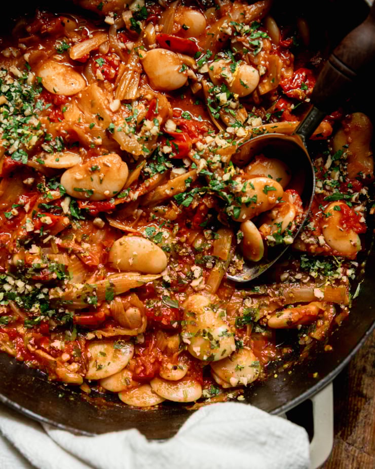 An up close, overhead shot shows a caramelized fennel, tomato and butter bean skillet garnished with chopped parsley and walnuts. A serving spoon is sticking out of the pan.