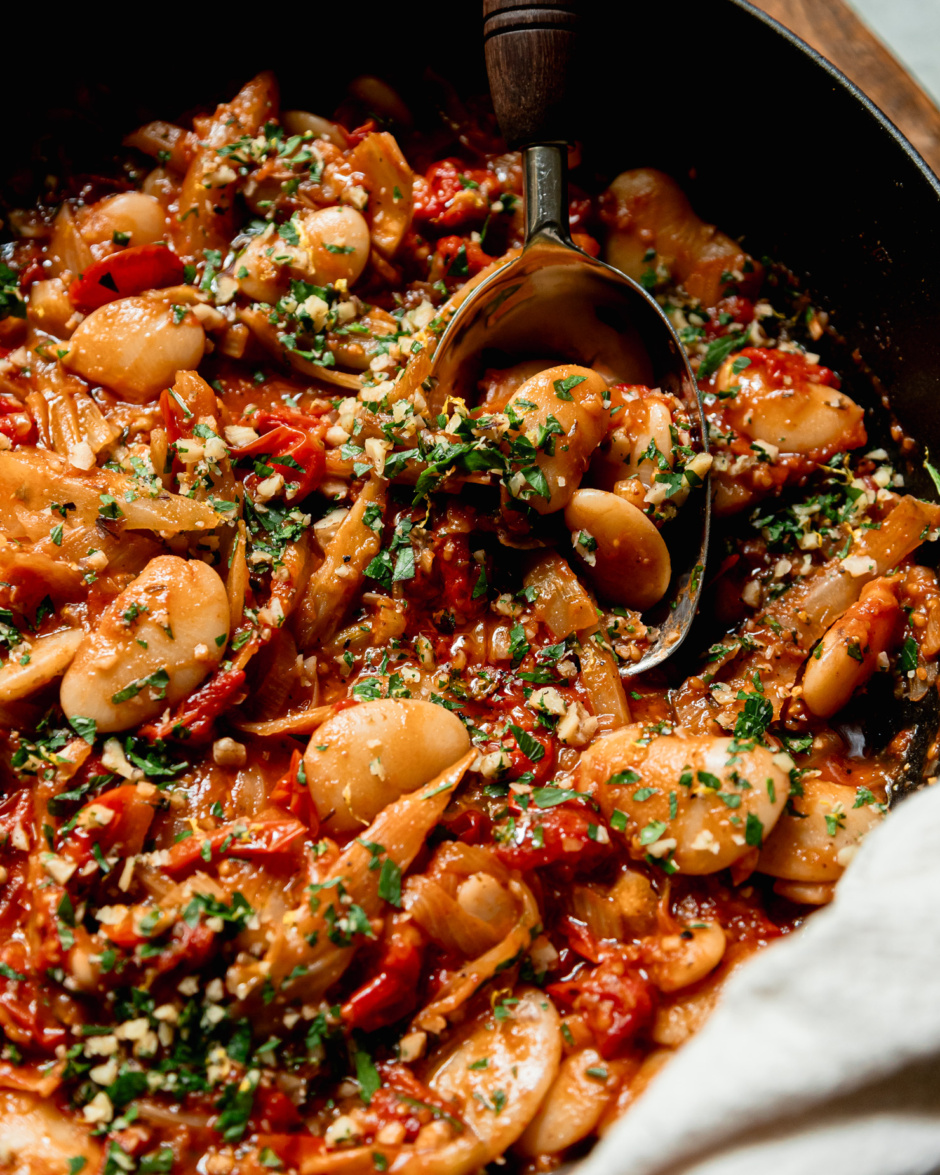 A close up, 3/4 angle shot shows a caramelized fennel, tomato and butter bean skillet garnished with chopped parsley and walnuts. A serving spoon is sticking out of the pan.