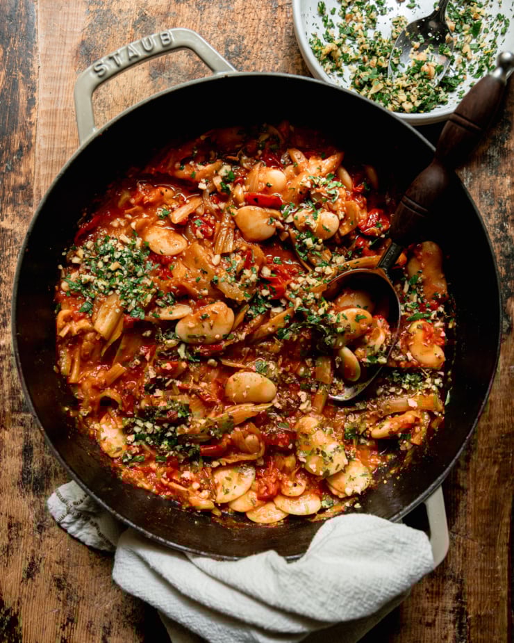 An overhead shot shows a caramelized fennel, tomato and butter bean skillet garnished with chopped parsley and walnuts. A serving spoon is sticking out of the pan.