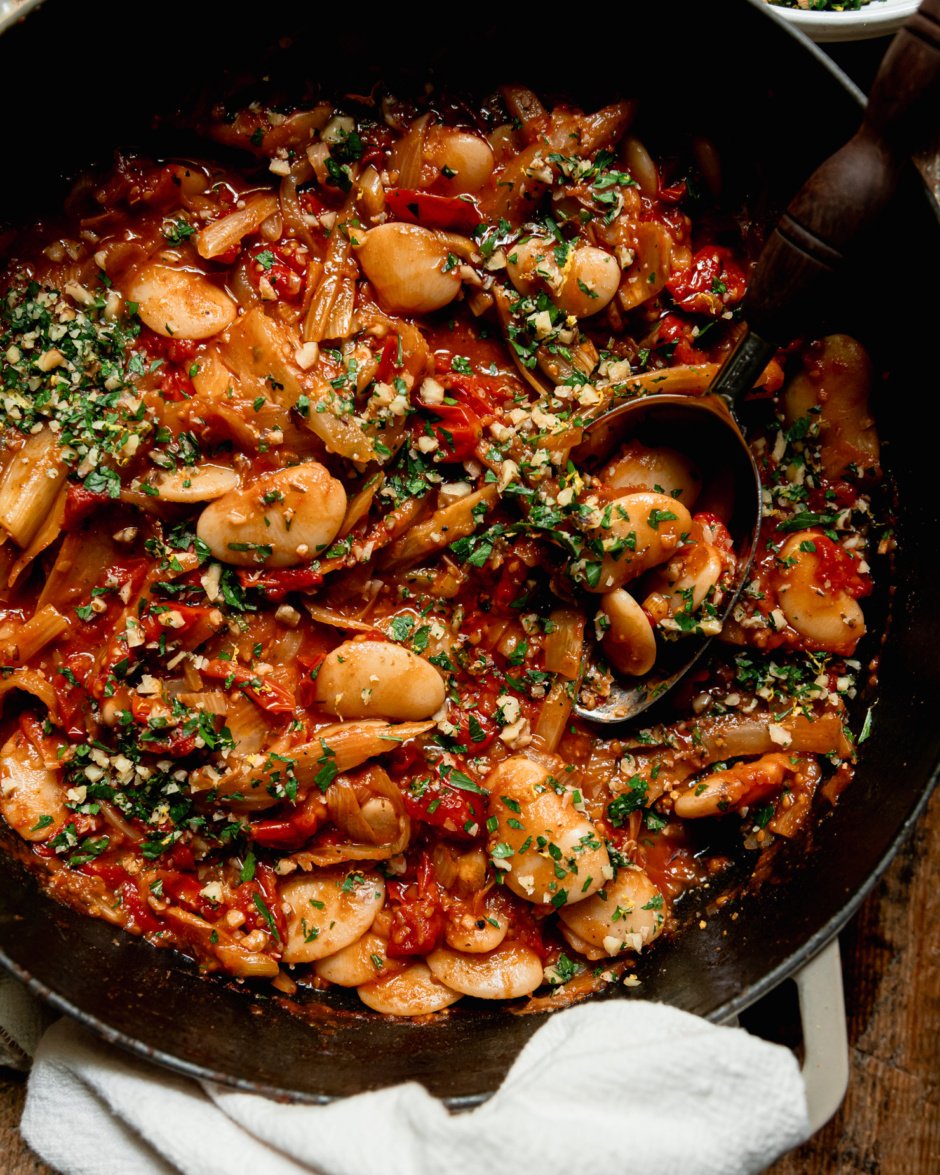 An up close, overhead shot shows a caramelized fennel, tomato and butter bean skillet garnished with chopped parsley and walnuts. A serving spoon is sticking out of the pan.