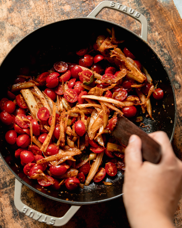 An overhead shot shows a hand stirring up some cooked fennel and cherry tomatoes in a brasier pot.