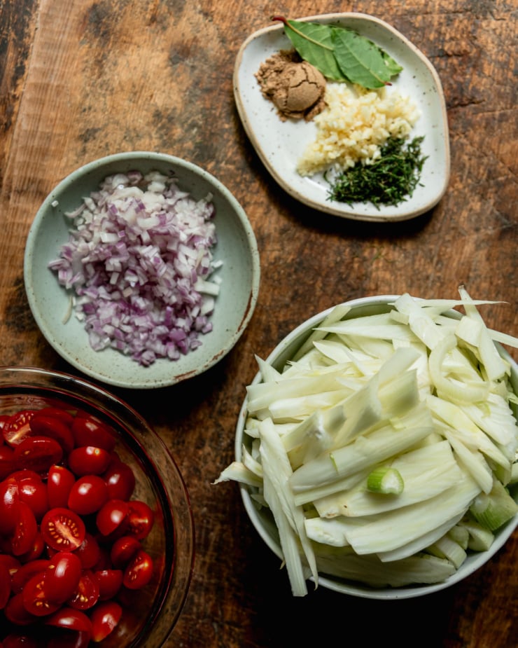 An overhead shot shows some prepped ingredients on a wooden board: minced garlic and thyme, sliced fennel, chopped shallots, and halved cherry tomatoes.