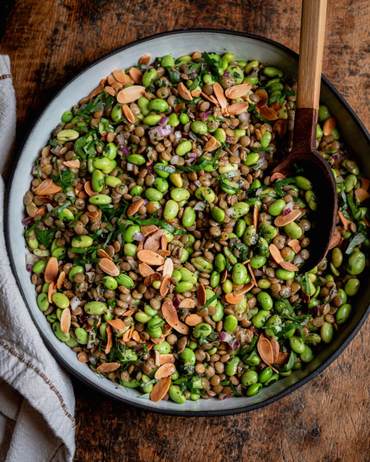 An overhead shot shows a green lentil edamame salad in a ceramic dish. The salad has chopped red onion, parsley, and a spiced lemon dressing, Toasted sliced almonds garnish the top and a wooden serving spoon is sticking out of the dish.