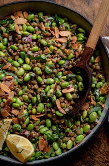 An overhead shot shows a green lentil edamame salad in a ceramic dish. The salad has chopped red onion, parsley, and a spiced lemon dressing, Toasted sliced almonds garnish the top and a wooden serving spoon is sticking out of the dish.