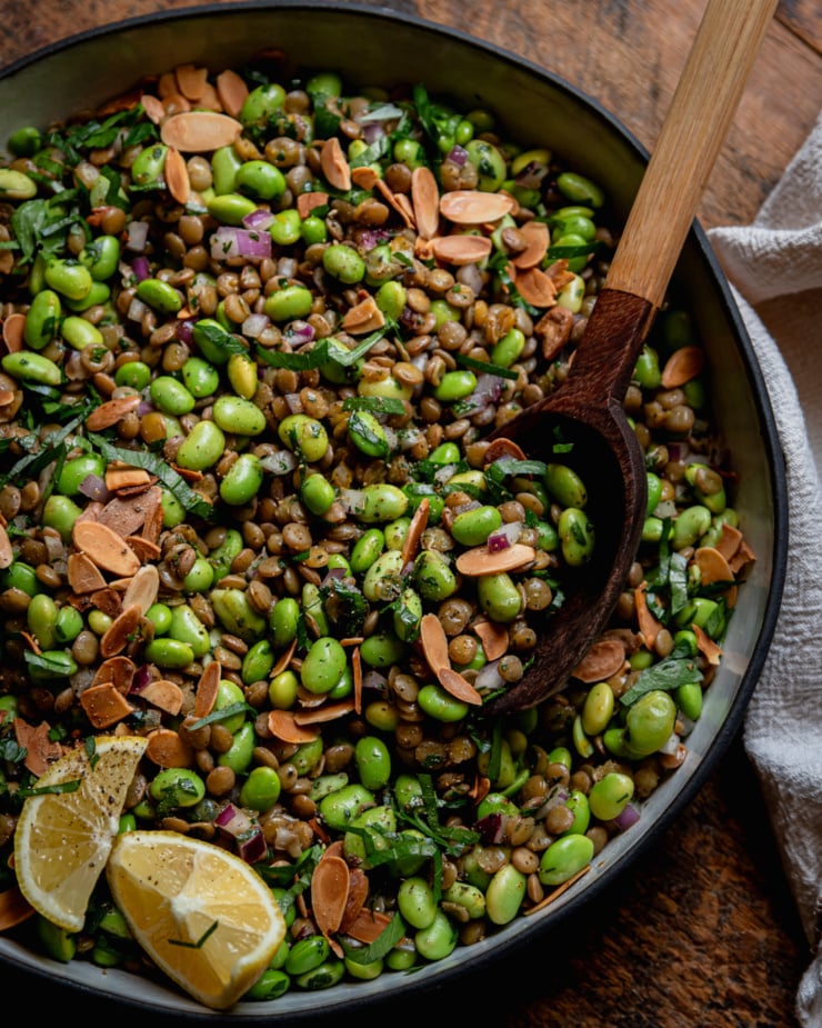 An overhead shot shows a green lentil edamame salad in a ceramic dish. The salad has chopped red onion, parsley, and a spiced lemon dressing, Toasted sliced almonds garnish the top and a wooden serving spoon is sticking out of the dish.