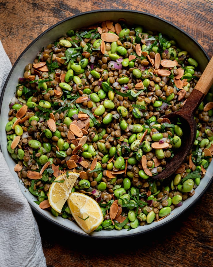 An overhead shot shows a green lentil edamame salad in a ceramic dish. The salad has chopped red onion, parsley, and a spiced lemon dressing, Toasted sliced almonds garnish the top and a wooden serving spoon is sticking out of the dish. Two lemon wedges are also perched on top of the salad.