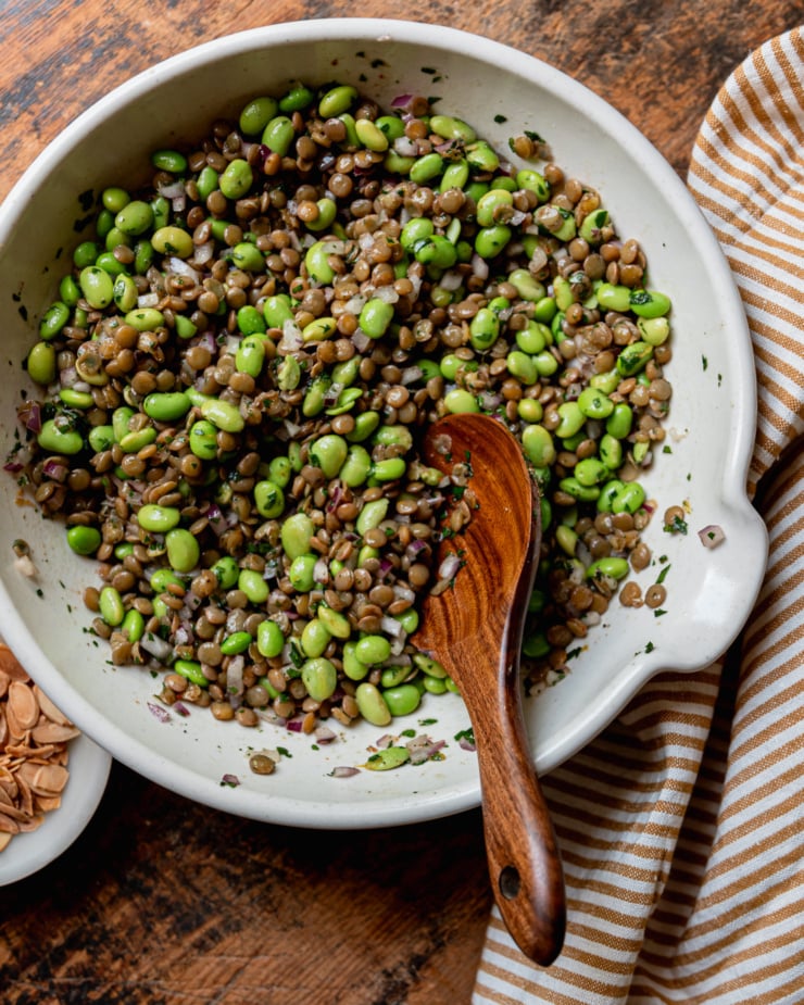 An overhead shot shows a green lentil edamame salad all mixed up in a ceramic bowl with a pouring spout. A small wooden spoon/paddle sticks out of the salad and a striped napkin is nearby.