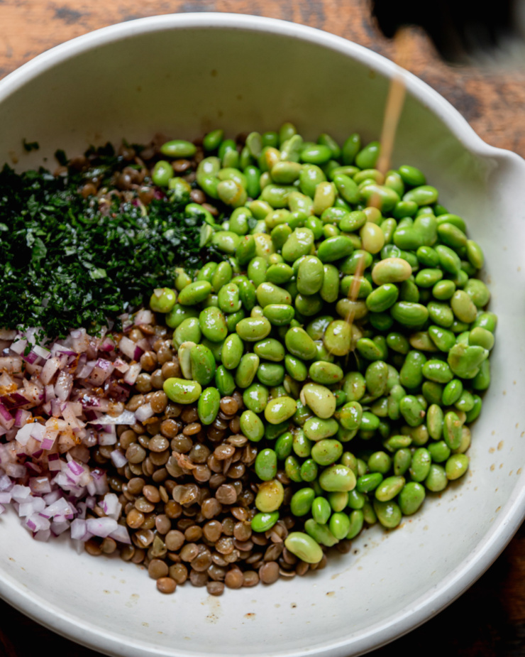 An overhead shot shows vinaigrette being poured over a bowl of thawed edamame, chopped parsley, cooked lentils, and chopped red onion.