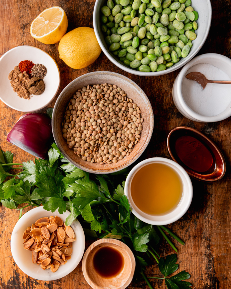 An overhead shot shows ingredients for a vegan salad: lemons, frozen shelled edamame, salt, maple syrup, avocado oil, dry green lentils, parsley, Worcestershire sauce, toasted sliced almonds, red onion, and spices.