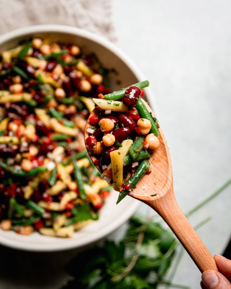 An overhead shot shows a hand holding a wooden spoon with a helping of four bean salad on top.