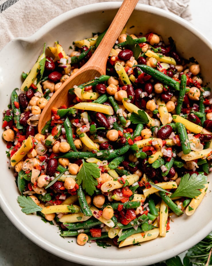 An overhead shot shows a large bowl filled with a tangy four bean salad. The salad features a rosemary red wine vinegar dressing, chopped roasted red peppers, parsley, and red onion. A spoon is sticking out of the salad bowl.