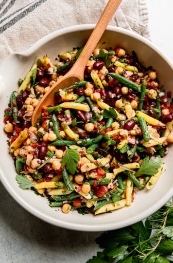 An overhead shot shows a large bowl filled with a tangy four bean salad. The salad features a rosemary red wine vinegar dressing, chopped roasted red peppers, parsley, and red onion. A spoon is sticking out of the salad bowl.