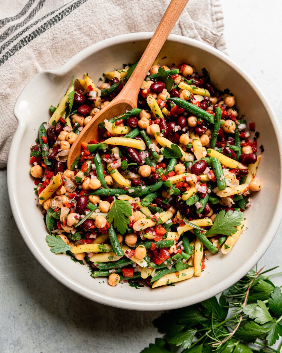 An overhead shot shows a large bowl filled with a tangy four bean salad. The salad features a rosemary red wine vinegar dressing, chopped roasted red peppers, parsley, and red onion. A spoon is sticking out of the salad bowl.