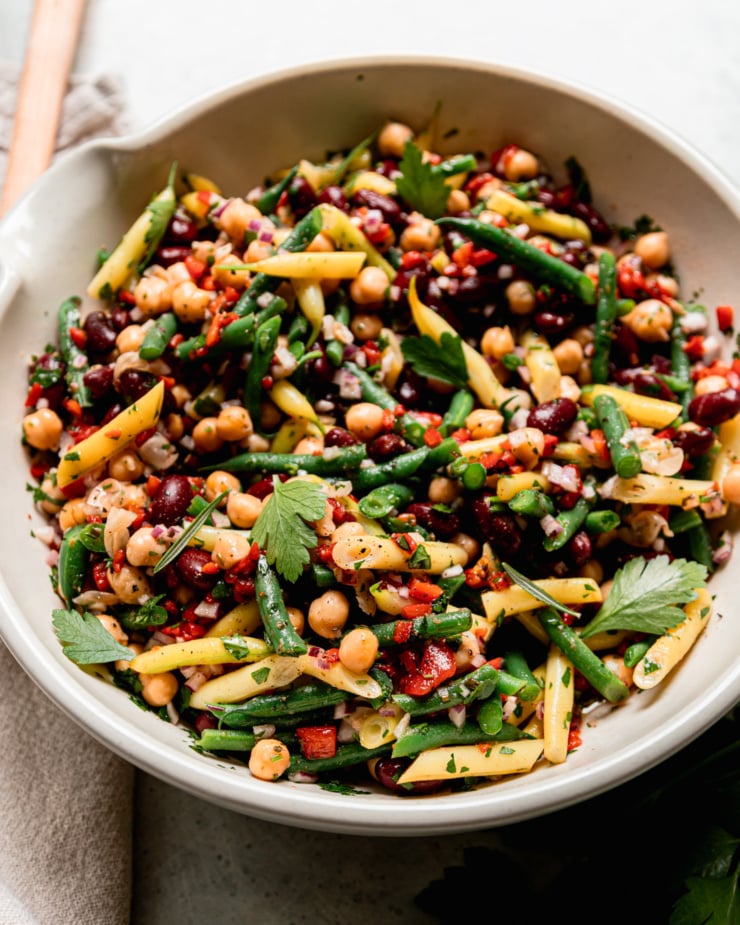 A 3/4 angle shot shows a large bowl filled with a tangy four bean salad. The salad features a rosemary red wine vinegar dressing, chopped roasted red peppers, parsley, and red onion.