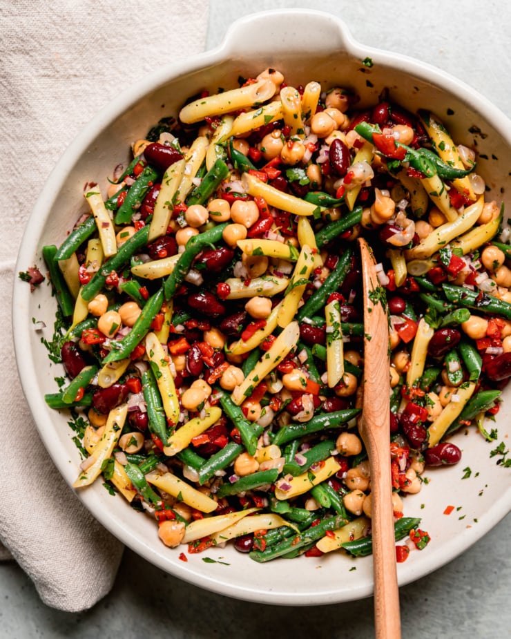 An overhead shot shows a large bowl filled with a tangy four bean salad. The salad features a rosemary red wine vinegar dressing, chopped roasted red peppers, parsley, and red onion. A spoon is sticking out of the salad bowl.