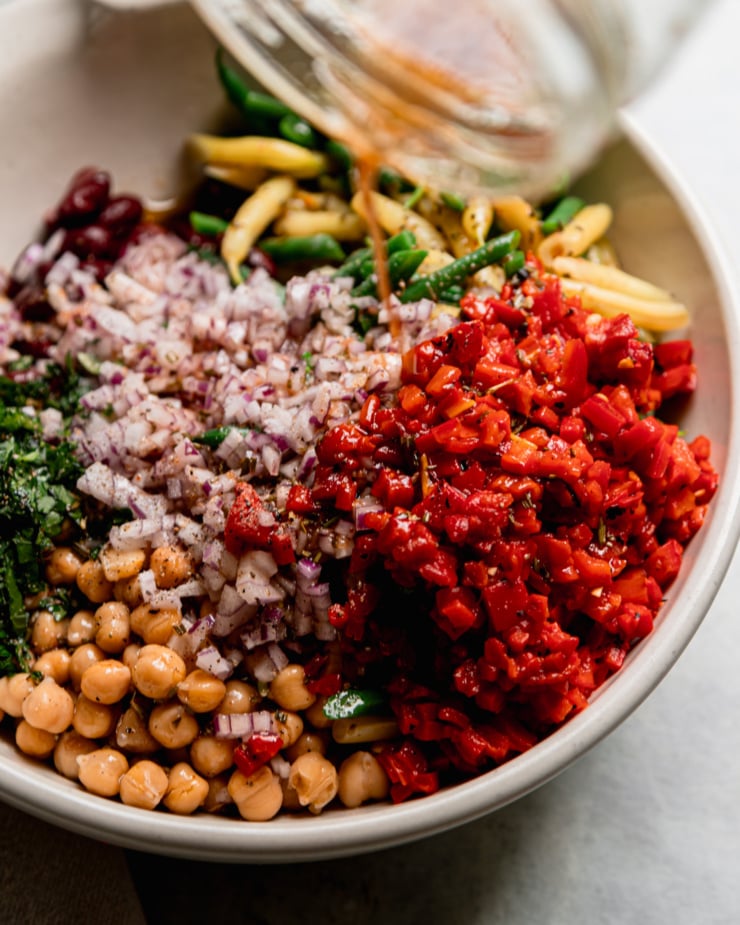 A 3/4 angle shot shows salad dressing being poured over a mixture of chopped roasted red peppers, chickpeas, chopped red onion, chopped parsley, red kidney beans, and cooked yellow/green beans.