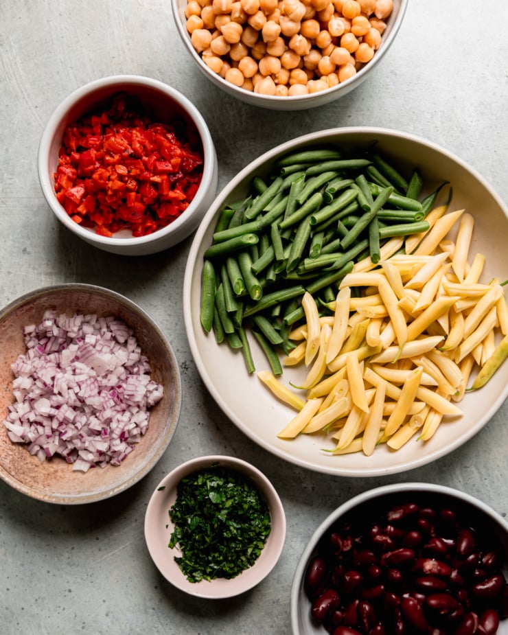 An overhead shot shows prepped ingredients: chopped green and yellow beans, chickpeas, red kidney beans, chopped parsley, chopped red onion, and chopped roasted red peppers. All ingredients are in bowls.