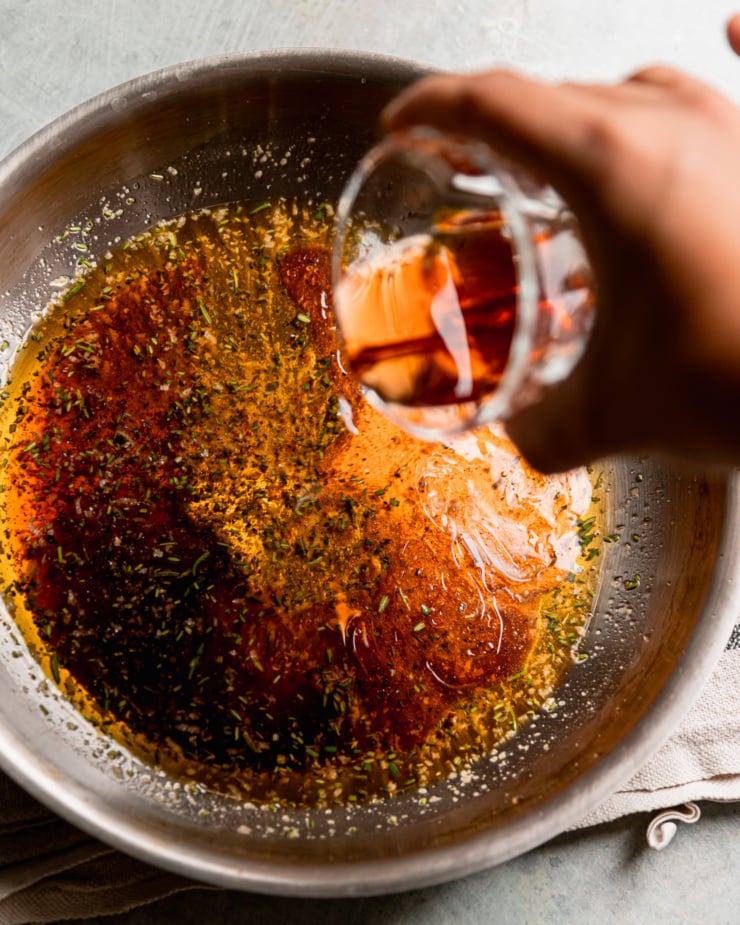 An overhead shot shows a hand pouring red wine vinegar into a sautรฉ pan with olive oil and spices.