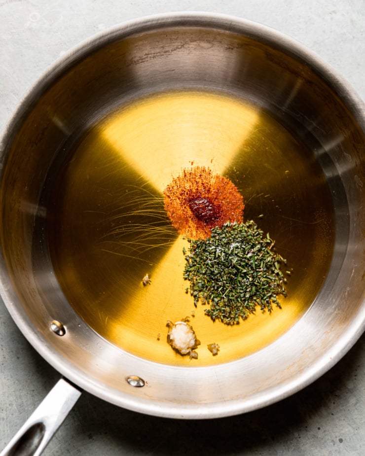 An overhead shot shows a steel sautรฉ pan with olive oil, garlic, rosemary, and chilies inside.