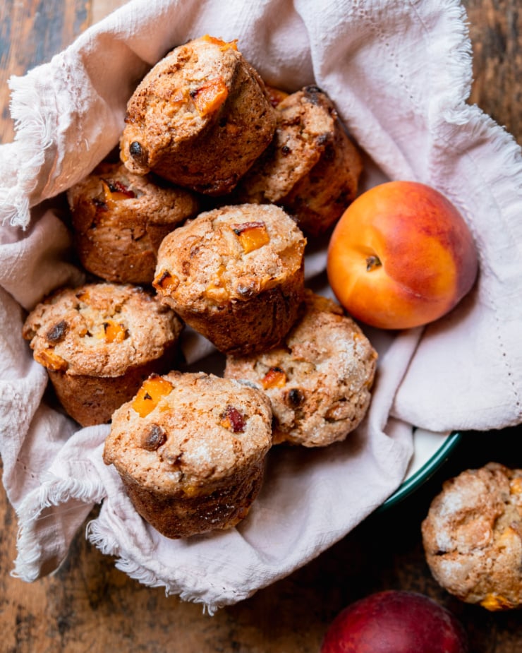 An overhead shot shows some vegan peaches and cream muffins nestled into a linen-lined bowl with a fresh peach in there too.