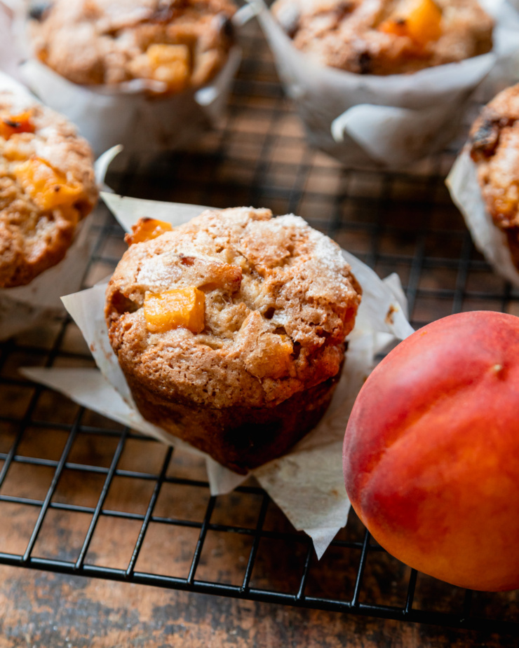 A 3/4 angle shot shows vegan peaches and cream muffins cooling in a wire rack against a rough wood board. A fresh peach is in the foreground.