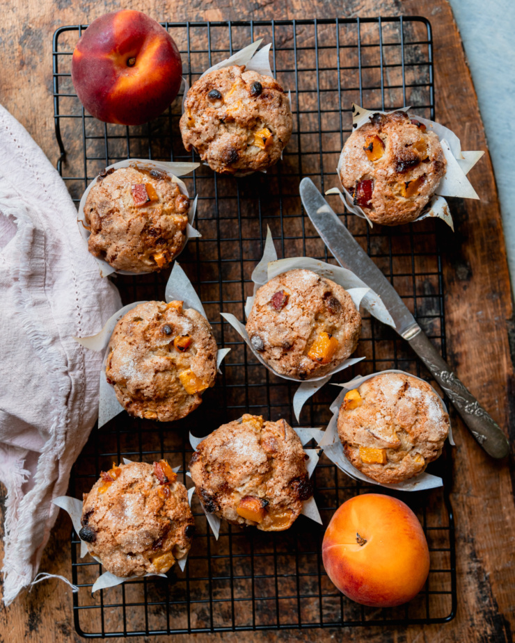 An overhead shot shows vegan peaches and cream muffins cooling on a wire rack. A couple fresh peaches are perched nearby, as well as a linen napkin.